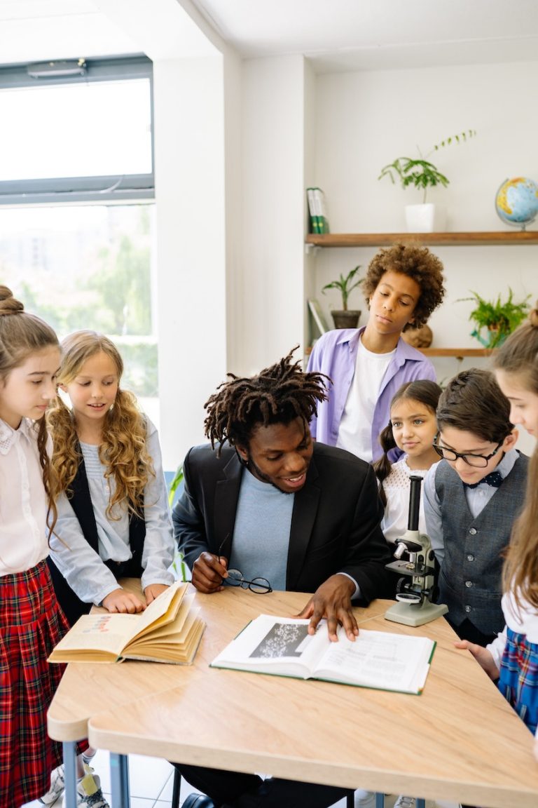 teacher standing around students showing something on a table